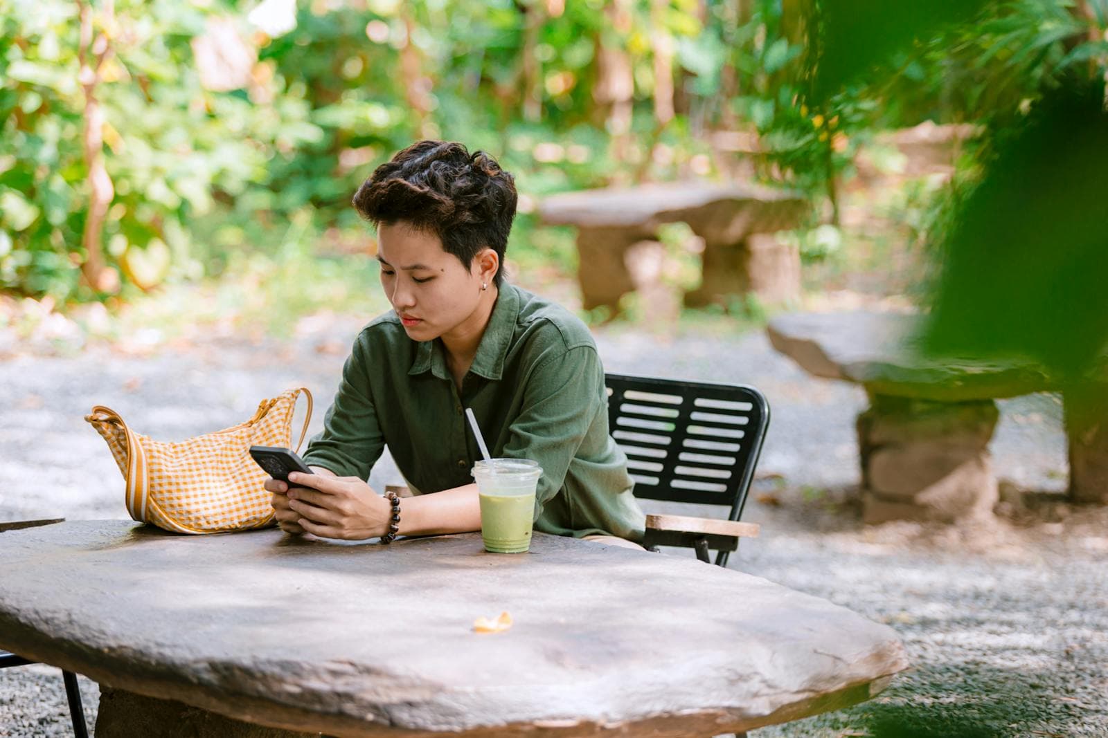Young adult relaxing outdoors at a stone table with a smartphone and a green smoothie, surrounded by lush greenery